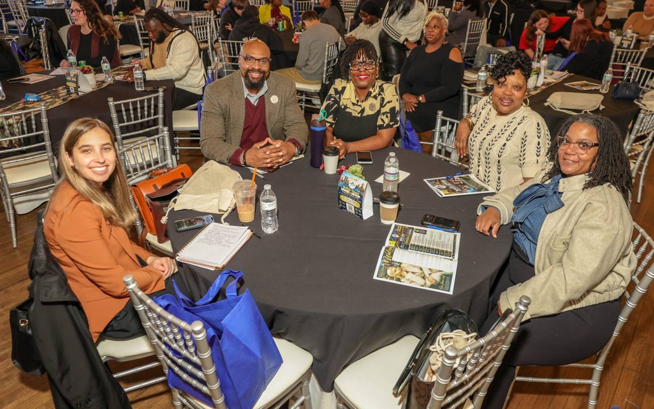 Business owners and partners smiling around a table at the 2026 Training Day Conference, with a full room behind them